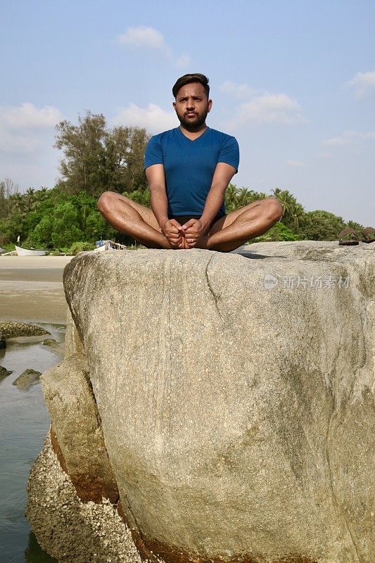 印度男子在岩石上练习蝴蝶/鞋匠姿势(Baddha Konasana)瑜伽姿势，水边，Palolem海滩，果阿，印度